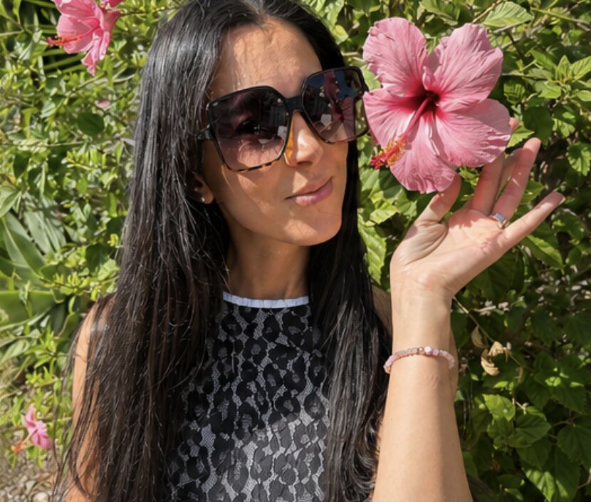 Junge Frau mit langen dunklen Haaren und großer dunkler Sonnenbrille hält eine pinkfarbene Hibiskusblüte vor grüner Hecke