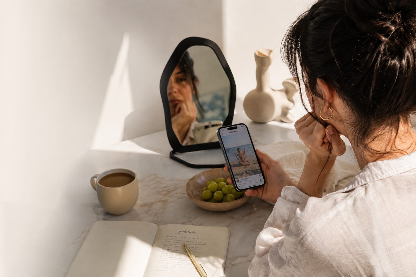 Frau am Schminktisch blickt nachdenklich auf Instagram am Smartphone, daneben Spiegel, Kaffee und ein Dankbarkeits-Journal